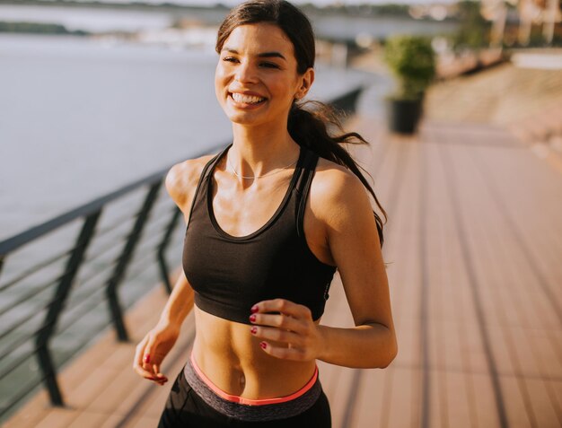 Mujer hermosa joven activa que corre en el paseo a lo largo de la orilla