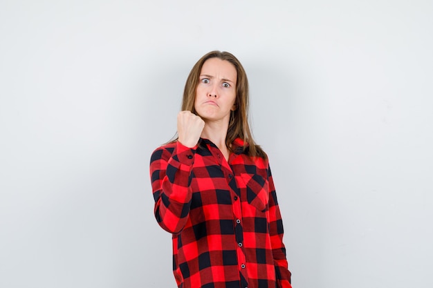 Mujer hermosa joven en advertencia de camisa casual con puño y mirando furioso, vista frontal.