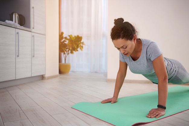 Mujer hermosa joven del ajuste que ejercita flexiones de brazos en una estera de la aptitud en casa en un día soleado. Concepto de entrenamiento en casa