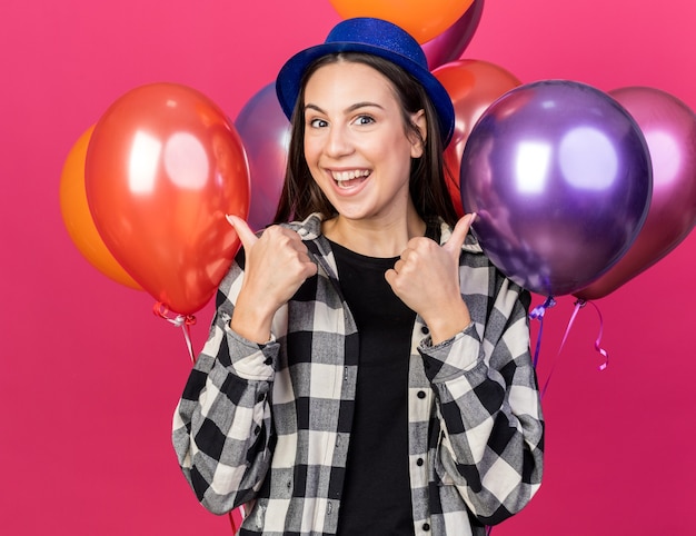 Mujer hermosa joven alegre que lleva el sombrero del partido que se coloca delante de los globos que muestran los pulgares para arriba aislados en la pared rosada