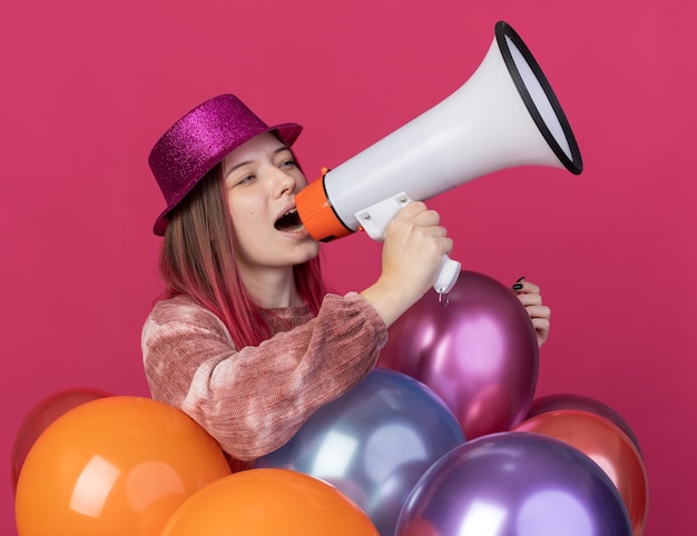 Mujer hermosa joven alegre que lleva el sombrero del partido que se coloca detrás de los globos que habla en el altavoz aislado en la pared rosada