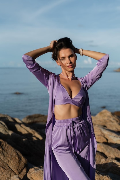 Mujer hermosa joven en un ambiente romántico ropa de seda lila en la playa contra el telón de fondo del mar y las piedras al atardecer