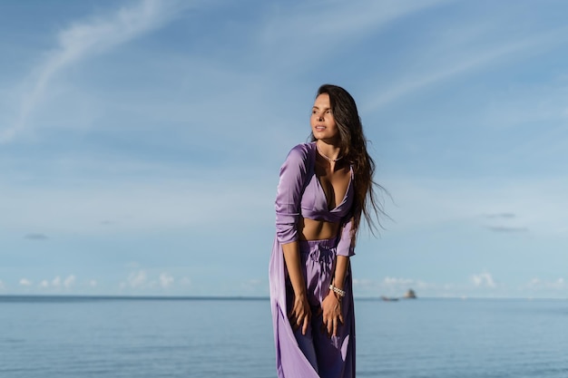 Mujer hermosa joven en un ambiente romántico ropa de seda lila en la playa contra el telón de fondo del mar y las piedras al atardecer
