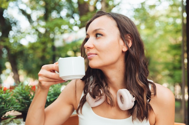 Una mujer hermosa joven con auriculares está sentada en una mesa en un café de verano y bebiendo café o té generación z