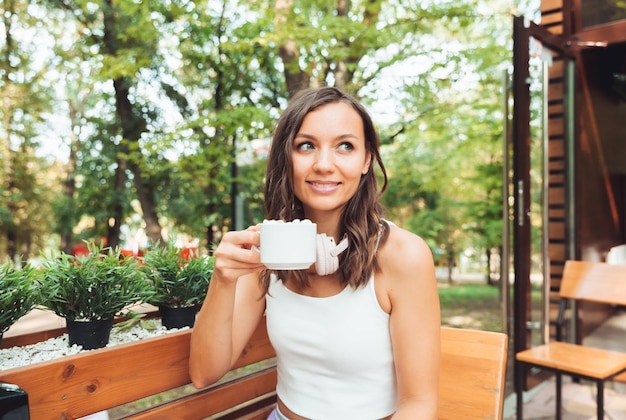 Una mujer hermosa joven con auriculares está sentada en una mesa en un café de verano y bebiendo café o té generación z