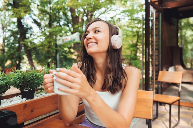 Una mujer hermosa joven con auriculares está sentada en una mesa en un café de verano y bebiendo café o té generación z