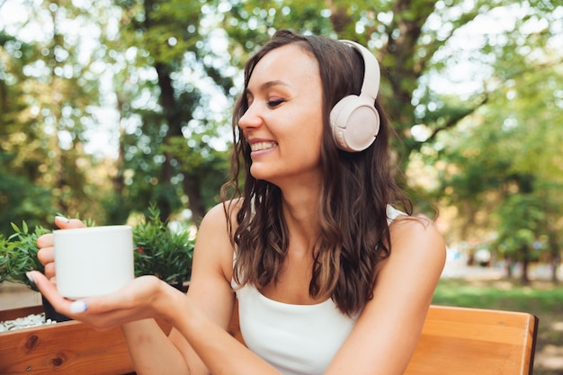 Una mujer hermosa joven con auriculares está sentada en una mesa en un café de verano y bebiendo café o té generación z