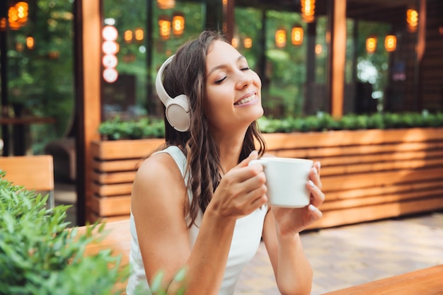 Una mujer hermosa joven con auriculares está sentada en una mesa en un café de verano y bebiendo café o té generación z