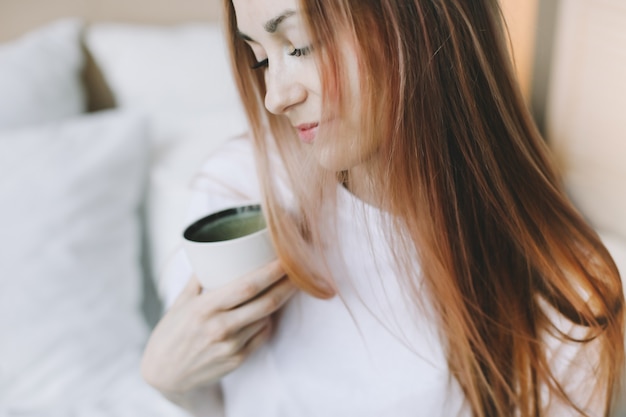 Mujer hermosa joven bebiendo té o café en la cama por la mañana en casa