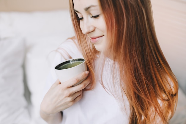 Mujer hermosa joven bebiendo té o café en la cama por la mañana en casa