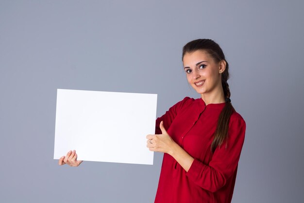 Mujer hermosa joven en blusa roja con trenza sosteniendo una hoja blanca de papel y mostrando el pulgar hacia arriba sobre fondo gris en el estudio