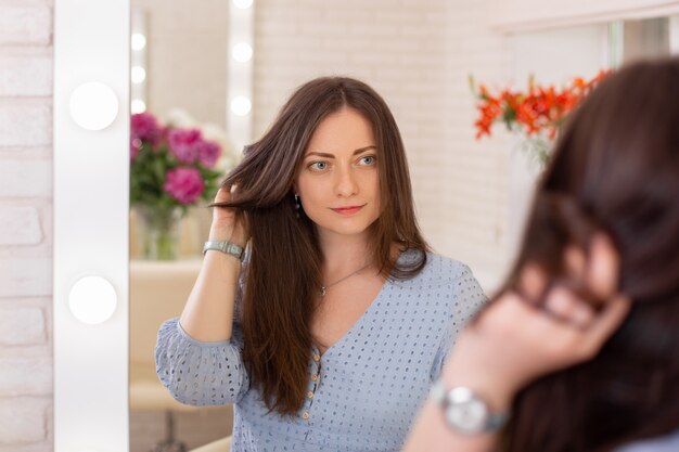 Mujer hermosa joven con cabello largo morena saludable mirando en el espejo en la peluquería