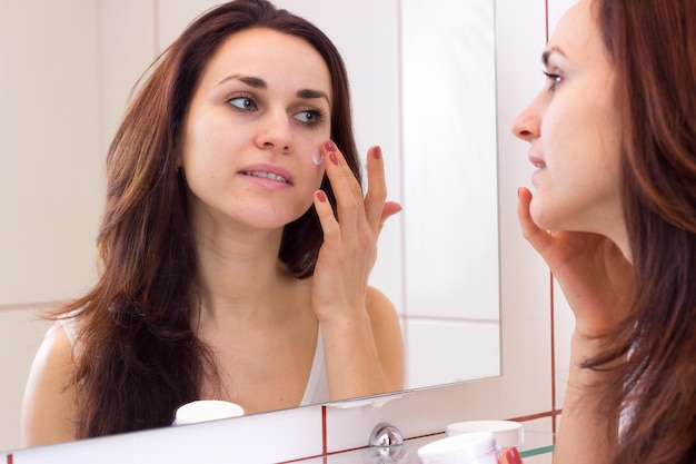 Mujer hermosa joven con cabello largo y oscuro en camisa blanca usando crema facial en su baño