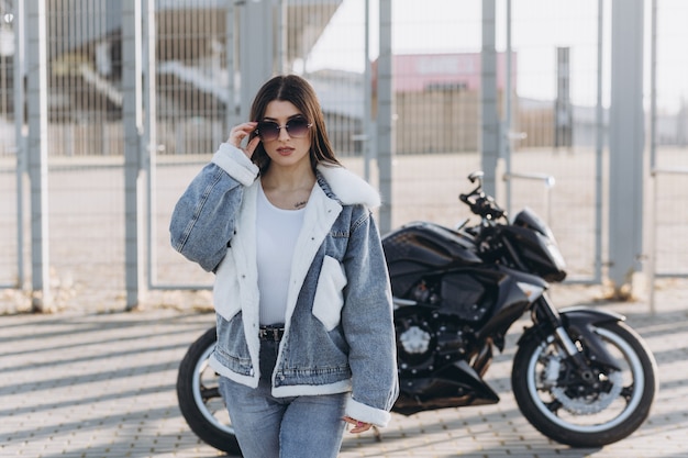 Mujer hermosa joven con cabello negro en pantalones de mezclilla y una chaqueta cerca de una motocicleta deportiva negra