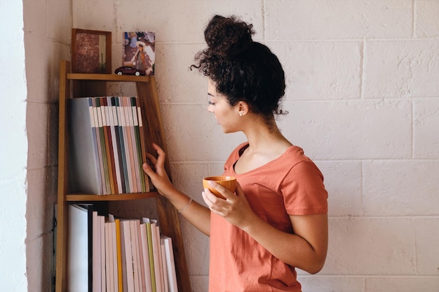 Mujer hermosa joven con cabello rizado oscuro en camiseta sosteniendo una taza de café en la mano eligiendo soñadoramente un libro de la estantería en un hogar acogedor