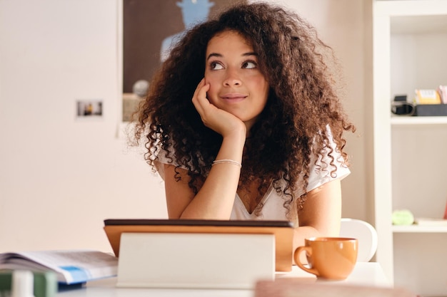 Mujer hermosa joven con cabello rizado oscuro sentada en la mesa con la tableta y la taza de café cerca de apoyarse en la mano mirando soñadoramente a un lado mientras pasa tiempo en casa