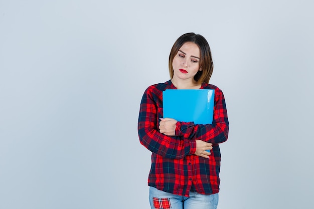 Mujer hermosa joven en camisa casual, jeans sosteniendo la carpeta, mirando hacia abajo y mirando molesto, vista frontal.