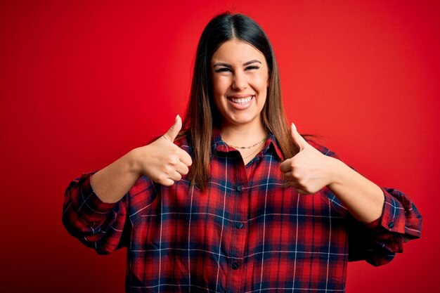 Foto mujer hermosa joven con camisa casual sobre fondo rojo signo de éxito haciendo gesto positivo con los pulgares hacia arriba sonriendo y feliz expresión alegre y gesto de ganador