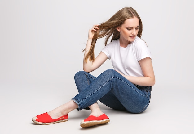 Mujer hermosa joven en una camiseta blanca y pantalones vaqueros sobre un fondo blanco. Mujer joven sentada sobre fondo blanco. Concepto de fotografía para anuncios de ropa y zapatos.