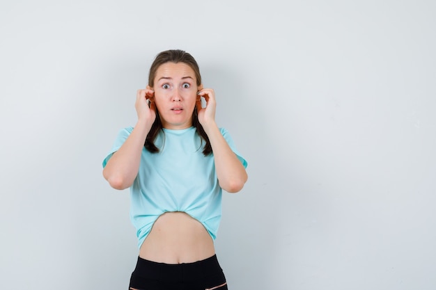 Mujer hermosa joven en camiseta con las manos en la cabeza y mirando asustado, vista frontal.