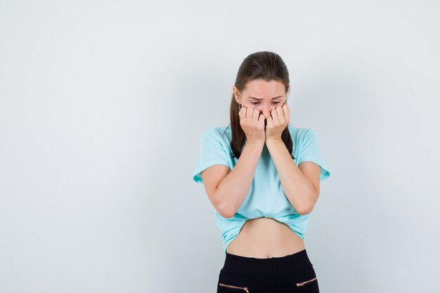 Mujer hermosa joven en camiseta, pantalones con las manos en la cara y mirando decepcionado, vista frontal.