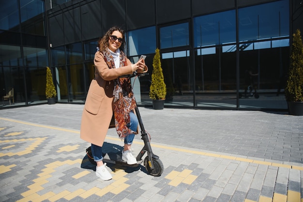 Mujer hermosa joven en una chaqueta sonríe y monta un scooter eléctrico para trabajar