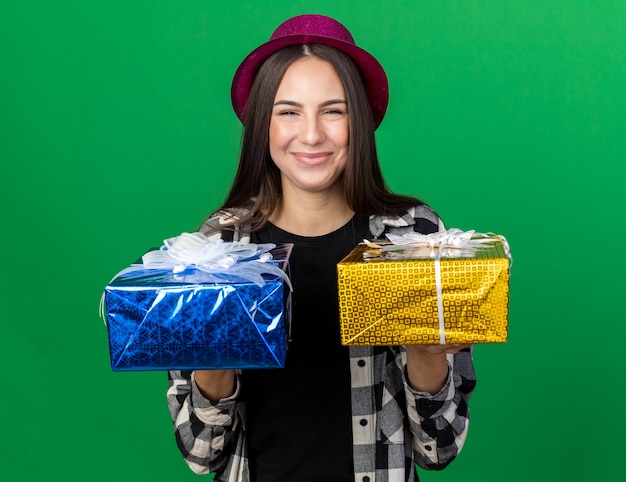 Mujer hermosa joven complacida con sombrero de fiesta sosteniendo cajas de regalo en la parte delantera aislada en la pared verde