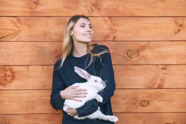 Mujer hermosa joven con un conejo blanco en sus brazos junto a una casa de madera en el campo