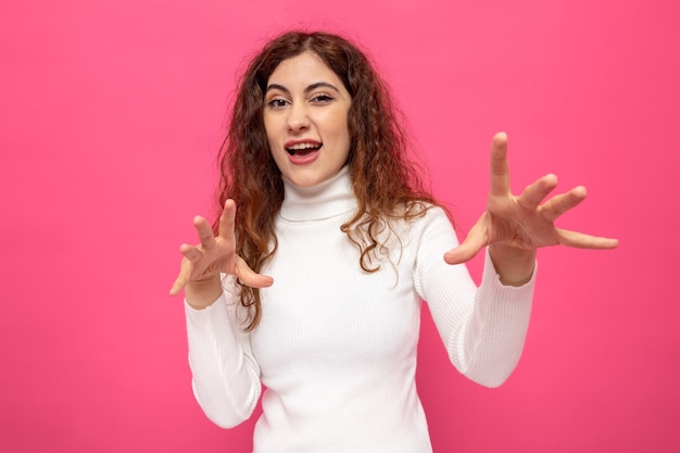 Mujer hermosa joven en cuello alto blanco feliz y alegre con las manos de pie sobre la pared rosa