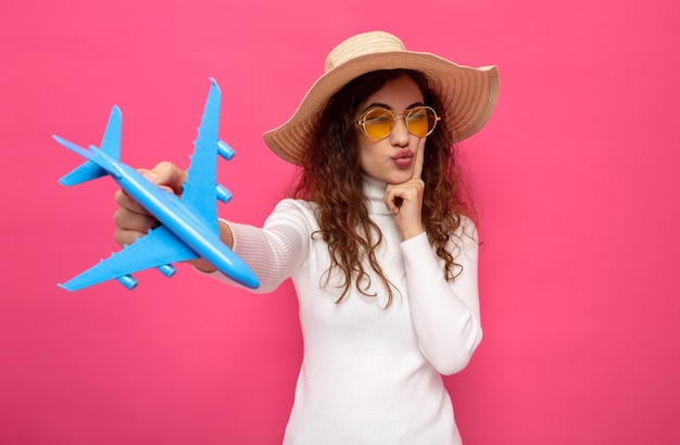 Mujer hermosa joven en cuello alto blanco con gafas amarillas y sombrero de verano con avión de juguete mirando a un lado con expresión pensativa pensando de pie en rosa