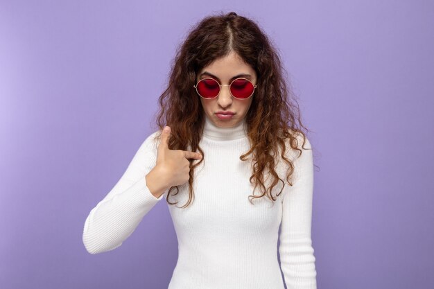 Mujer hermosa joven en cuello alto blanco con gafas rojas mirando hacia abajo con expresión triste apuntando a sí misma con el dedo índice