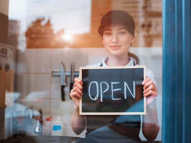 Mujer hermosa joven en un delantal un trabajador de café tiene un cartel abierto con el telón de fondo de un bistro detrás de la ventana de cristal