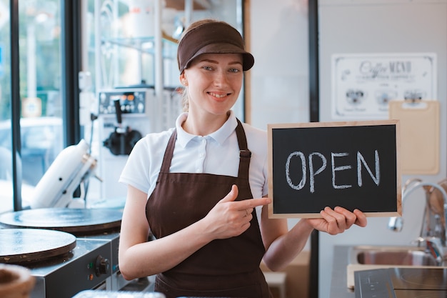 Mujer hermosa joven en un delantal un trabajador de café tiene un cartel abierto con el telón de fondo de un bistro