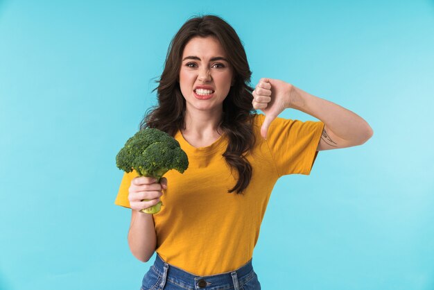 mujer hermosa joven disgustada posando aislada sobre pared azul sosteniendo brócoli mostrando los pulgares hacia abajo.