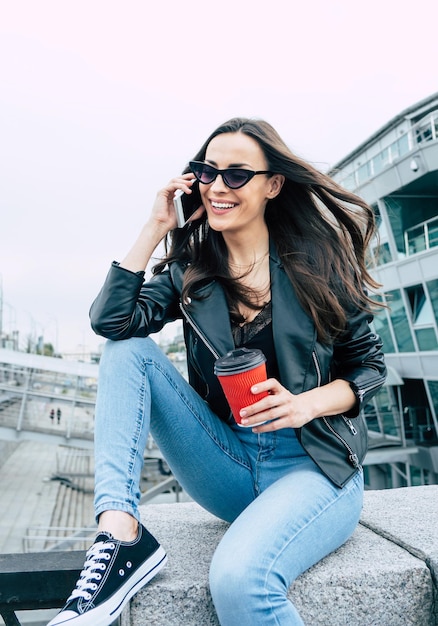 Mujer hermosa joven elegante y de moda con gafas de sol y estilo hipster mientras usa un teléfono y se divierte al aire libre en la ciudad