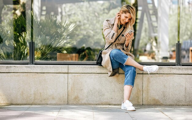 Mujer hermosa joven escribiendo mensajes en el teléfono inteligente sentado al aire libre en la calle de la ciudad en un día soleado Mujer bonita usando conexión inalámbrica gratuita en el teléfono celular Concepto de viaje y tecnología