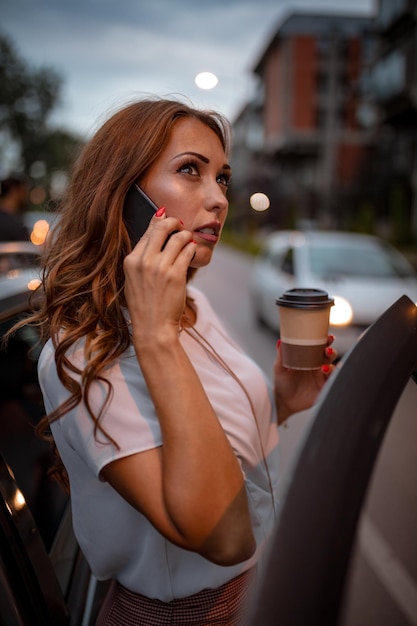 Una mujer hermosa joven está hablando en el teléfono inteligente con una taza de café y va a entrar en el coche.