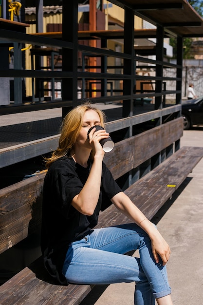 Una mujer hermosa joven está sentada en un banco y bebiendo café