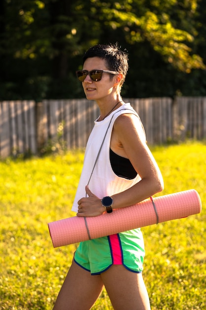 Mujer hermosa joven con estera de yoga en un parque, pelo negro corto, gafas de sol y pantalones cortos, deporte al aire libre en verano, chica fitness delgada en ropa deportiva, mujer en entrenamiento de entrenamiento