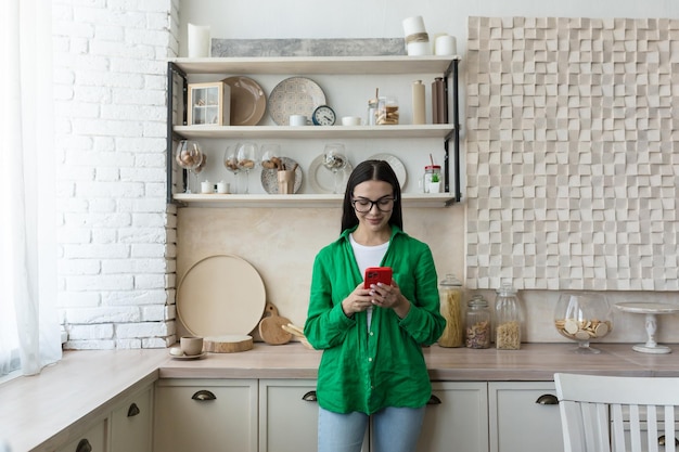 Mujer hermosa joven con gafas y camisa verde de pie en casa en la cocina escribiendo mensajes en