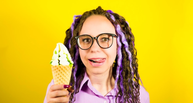 Mujer hermosa joven en gafas lamiendo labios sobre fondo amarillo Retrato de mujer feliz con rastas en anticipación disfrutando de sundae