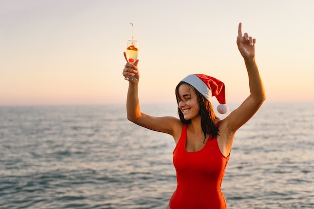 Mujer hermosa joven en un gorro de Papá Noel con una copa de champán en sus manos caminar por la playa