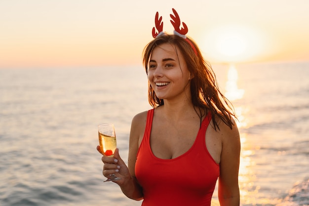 Mujer hermosa joven en un gorro de Papá Noel con una copa de champán en sus manos caminar por la playa