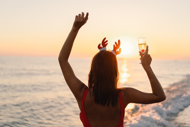 Mujer hermosa joven en un gorro de Papá Noel con una copa de champán en sus manos caminar por la playa