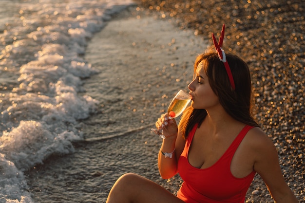 Mujer hermosa joven en un gorro de Papá Noel con una copa de champán en sus manos caminar por la playa