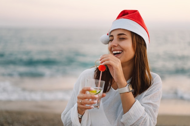 Mujer hermosa joven en un gorro de Papá Noel con una copa de cóctel en sus manos caminar por la playa