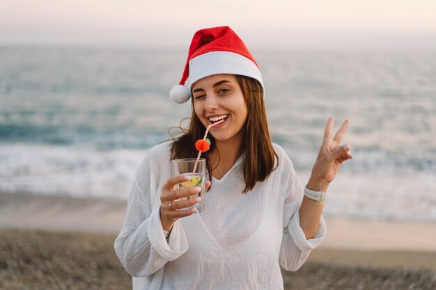Mujer hermosa joven en un gorro de Papá Noel con una copa de cóctel en sus manos caminar por la playa