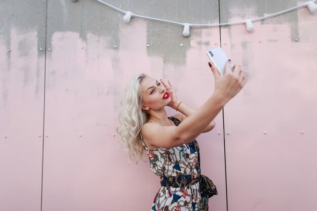 Mujer hermosa joven inconformista en vestido de moda haciendo selfie en teléfono junto a la pared rosa. Chica blogger hace la transmisión