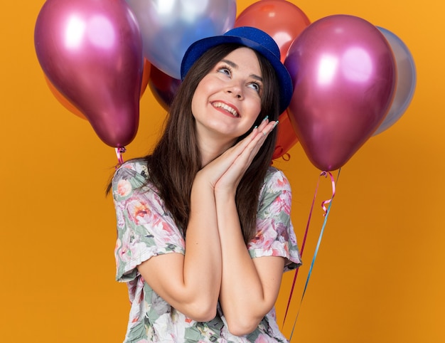 Mujer hermosa joven de lado mirando complacido con sombrero de fiesta de pie delante de globos aislados en la pared naranja