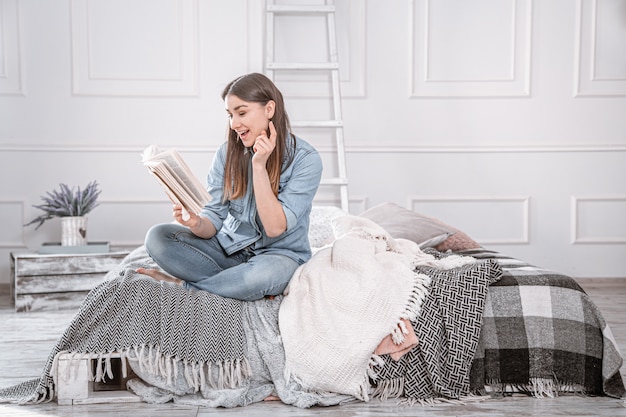 Mujer hermosa joven leyendo un libro en la cama en el interior de una habitación acogedora.